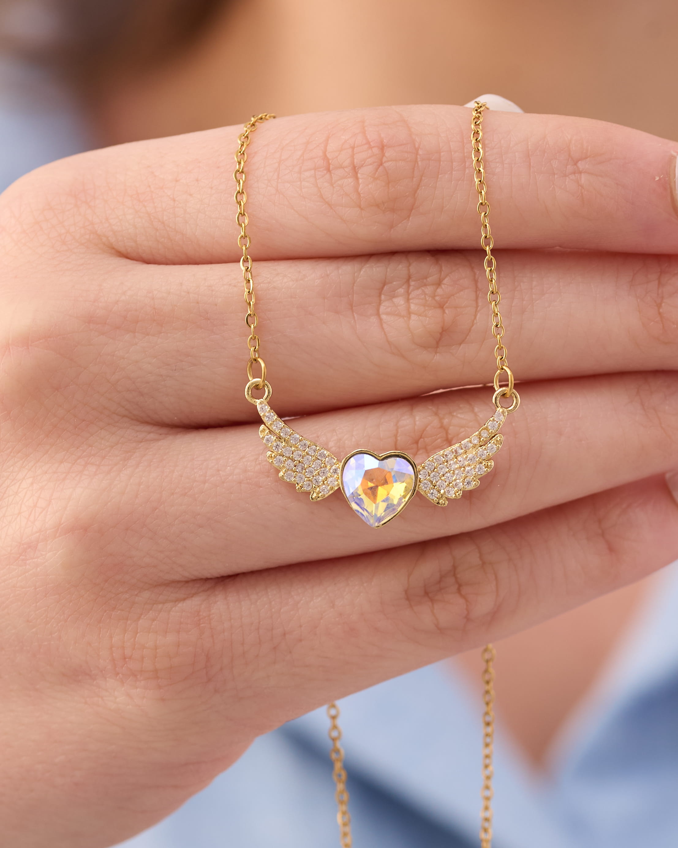 Stainless steel gold-tone angel wings necklace with iridescent crystal heart pendant held in hand
