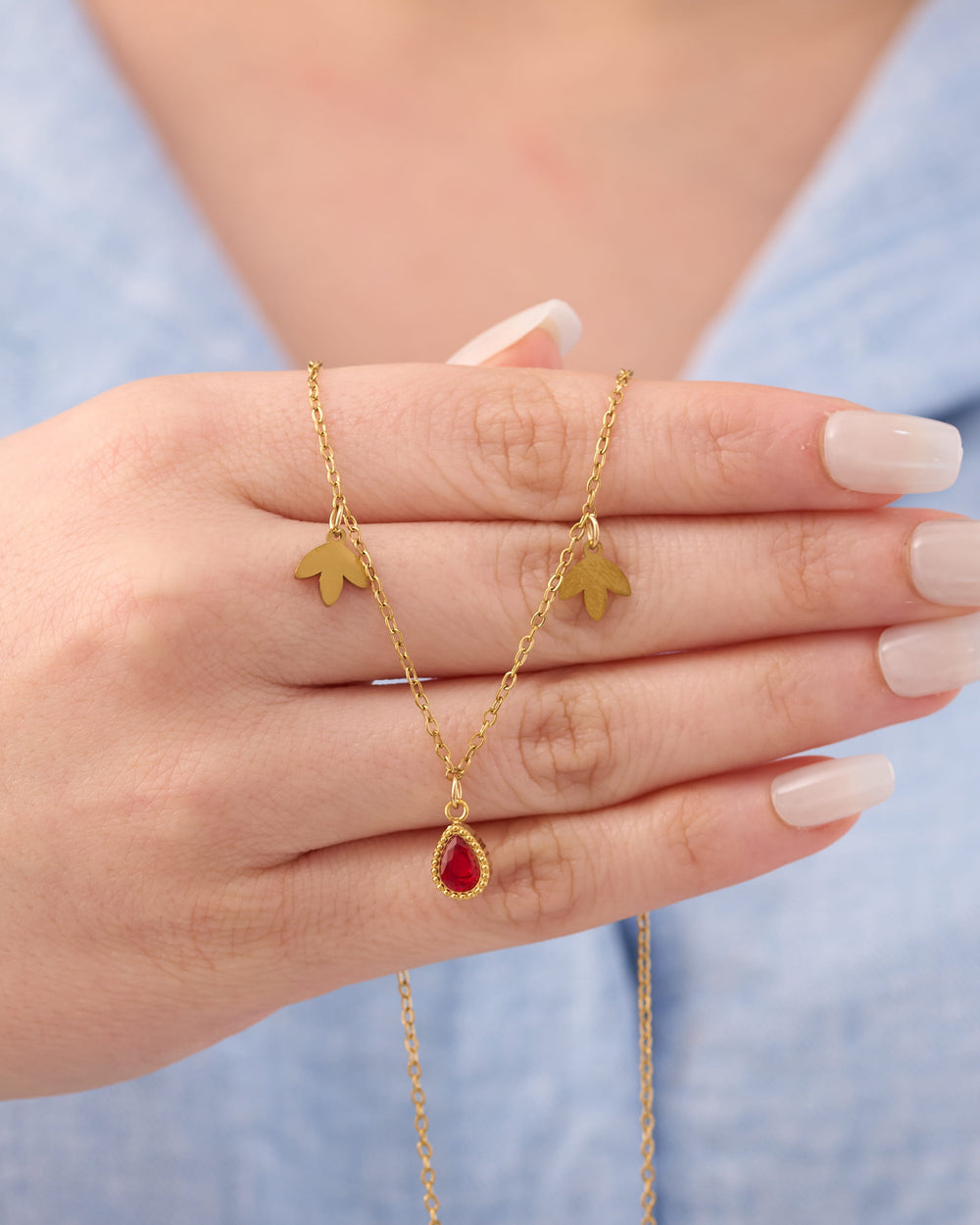 Stainless steel gold-tone charm necklace with maple leaf and red teardrop crystal pendant held in hand